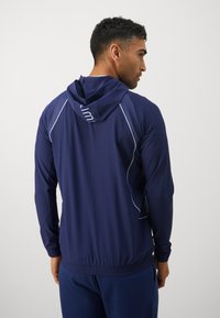 Tums zilgs sporta jaka ar kapuci, ar acs auduma akcentiem, kontrastējošu šuvju un elastīgu jostasvietu. Gluda auduma un pielāgota dizaina.