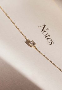 Bracelet en chaîne dorée présentant un cristal rond central, monté sur un fermoir à quatre griffes, reposant sur une page de note beige texturée.