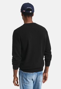 Maglione nero con maniche lunghe, scollatura rotonda e polsini a coste. Indossato sopra jeans azzurri; persona di spalle rispetto alla fotocamera.