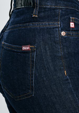 Prim-plan al buzunarului din spate al unei perechi de blugi din denim albastru închis, cu cusături portocalii și o etichetă roșie cu inscripția „BIG STAR” pe partea dreaptă.