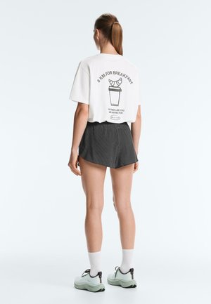 Žena s copom nosi biele tričko s textom „8 KM FOR BREAKFAST“ a grafikou croissantu a šálky kávy, čierne šortky, biele ponožky a tenisky.