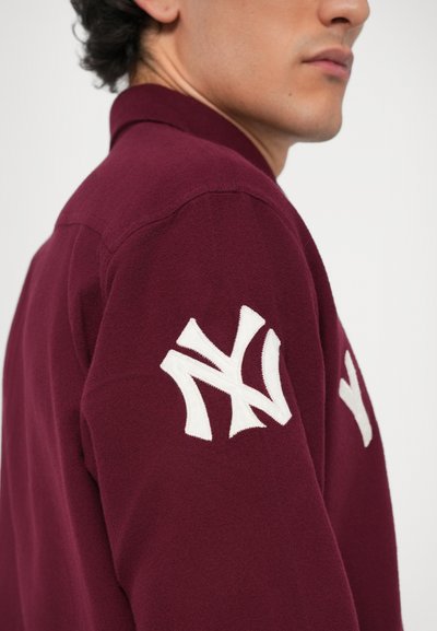Bordeaux jakke med krage, som har en stor hvit brodert New York Yankees-logo på venstre erme og et glatt teksturert stoff.