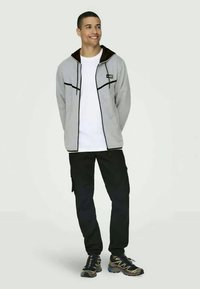 Graue Zip-Jacke mit schwarzen Akzenten, kombiniert mit einem weißen T-Shirt und schwarzen Cargo-Hosen. Bemerkenswerte Merkmale sind eine mit braunem Stoff gefütterte Kapuze und sportliche Schuhe.