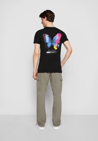 Black cotton t-shirt featuring a colorful butterfly graphic on the back. Paired with light green cargo pants. Simple design and casual fit.