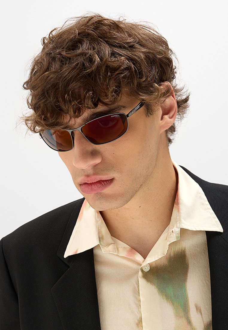 Lunettes de soleil avec des verres teintés et un design légèrement enveloppant. Présentent des montures noires fines et des détails matériels minimes, portées avec une chemise à motifs.