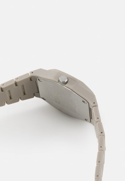 Quarzuhr mit einem beigen Kunststoffband und -gehäuse. Die metallisch silberne Rückseite ist sichtbar und verfügt über eine hexagonale Krone sowie minimale Details am Band.