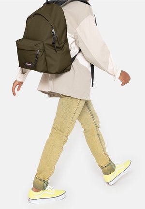 Personne marchant portant des baskets jaunes, une veste beige clair, un jean délavé, et un sac à dos vert olive avec une poche avant.