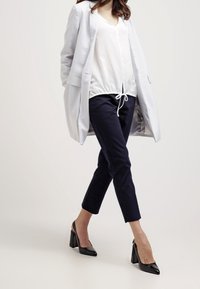 Světlý šedý blejzr přes bílou blůzu se stahovací šňůrkou, tmavě modré kalhoty a černé lodičky na podpatku. Oblečení má čistý a moderní design.