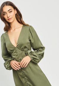 Olivno zelena satenasta obleka z globokim V-izrezom, napihnjenimi dolgimi rokavi in dekorativnim gumbom ob sprednji strani. Gladka tekstura in prilagojen pas.