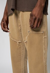 Närbild av en persons händer som vilar på beige byxor med unik sömnad och fransade kanter, som visar dekorativa fickor och knappar.