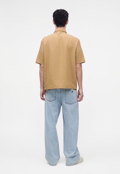 Homme de dos portant une chemise beige à manches courtes et un jean bleu clair à jambes larges sur fond blanc uni.