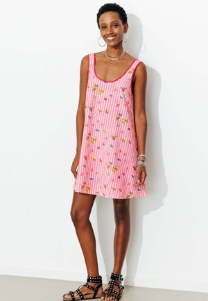Femme souriante aux cheveux courts portant une robe rose sans manches à imprimé floral et des sandales noires cloutées, debout contre un mur blanc.