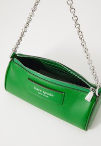 Zielona skórzana torebka o cylindrycznym kształcie, z srebrnym łańcuszkiem, z zamkiem błyskawicznym oraz naszywką z logo "kate spade NEW YORK". Wnętrze widoczne.