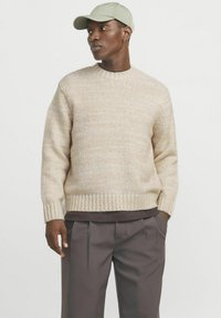 Beige knit sweater with a round neckline and ribbed cuffs, paired with dark pleated trousers and a light green baseball cap.
