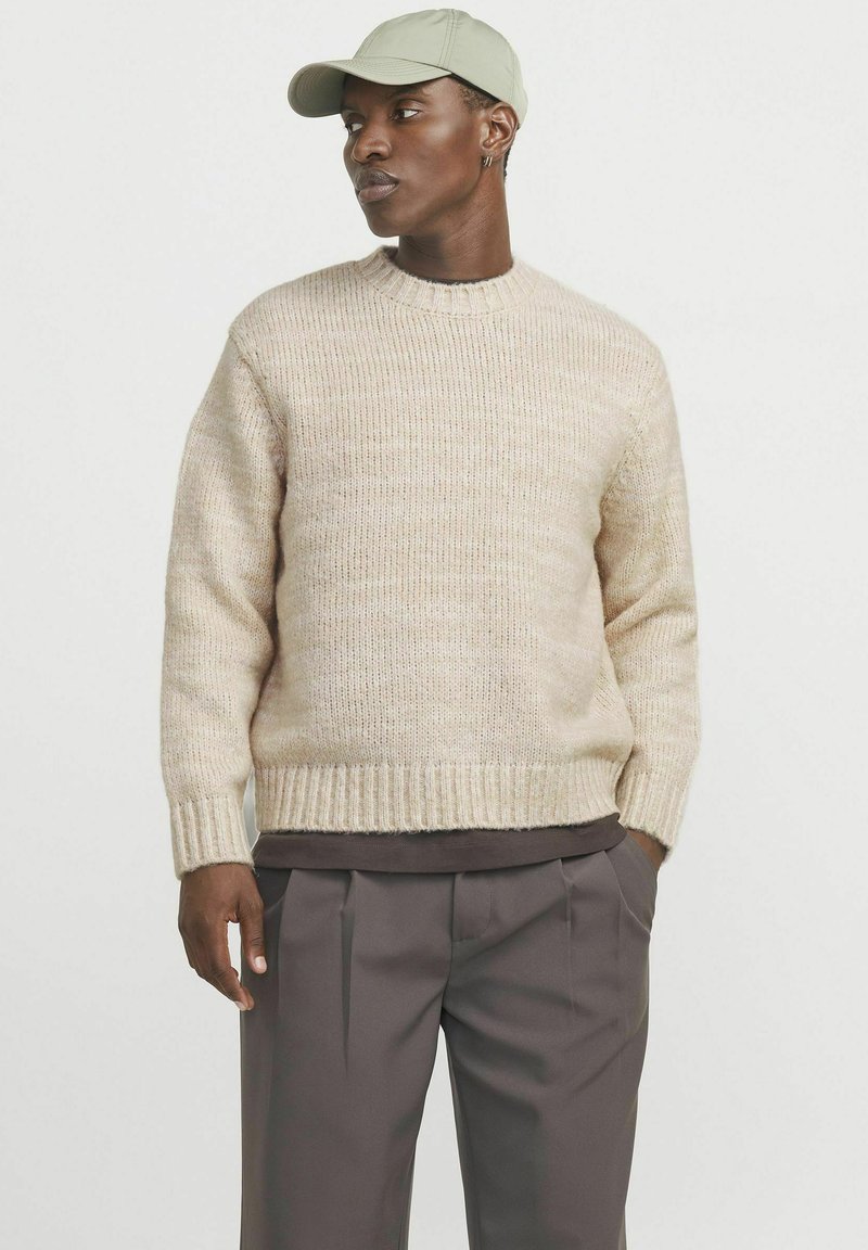 Beige Strickpullover mit rundem Ausschnitt und gerippten Bündchen, kombiniert mit dunklen, plissierten Hosen und einer hellgrünen Baseballmütze.