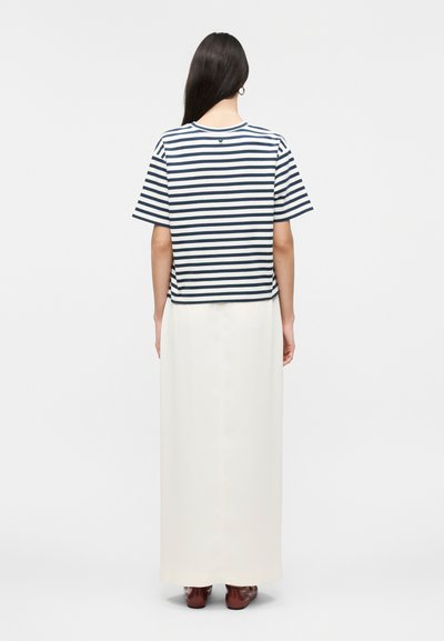 Γυναίκα με μακριά σκουρόχρωμα μαλλιά, που φοράει μπλε σκούρα και λευκή ριγέ κοντομάνικη μπλούζα, λευκή μακριά φούστα και καφέ παπούτσια, γυρισμένη πλάτη σε απλό φόντο.