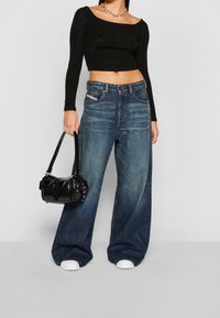 Musta pitkähihainen lyhyt toppi, jossa on avara kaula-aukko, yhdistettynä leveälahkeisiin sinisiin denimi-farkkuihin, joissa on haalistunut pesu, ja mustaan käsilaukkuun.