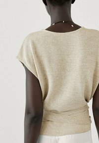 Beige knit top with short sleeves, relaxed fit, and a textured finish. Features a wrap detail at the waist. Background is neutral.