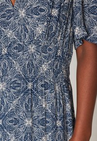 Gros plan sur un vêtement bleu marine orné de motifs floraux blancs complexes et de tissu froncé à la taille, porté par une personne à la peau foncée.