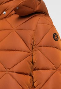 Polstret jakke i varm orange med et diamantquiltet mønster, der har hætte, trykknapdetaljer og et rundt logo-patch på ærmet.