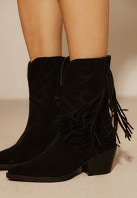 Μαύρες σουέτ μπότες μέχρι τη μέση της γάμπας με ραφές σε δυτικό στιλ και λεπτομέρεια με κρόσσια, φορεμένες σε ανοιχτόχρωμα πόδια, που στέκονται σε μπεζ πάτωμα.