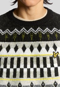 Πλεκτό μάλλινο πουλόβερ με μαύρη βάση, γεωμετρικά λευκά και γκρι σχέδια, κίτρινες λεπτομέρειες και μικρό κεντητό κίτρινο λογότυπο στο κάτω δεξί μέρος.