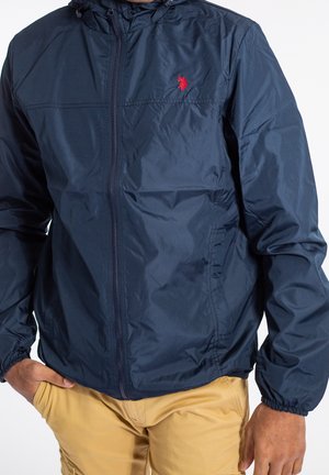 Mand iført en navyblå lynlåstrøje med lille rødt logo og beige bukser, den ene hånd i lommen, vist fra skulder til talje.