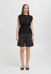 Top nero plissettato senza maniche con vita arricciata, abbinato a pantaloni corti neri a vita alta e ballerine nere su uno sfondo neutro.