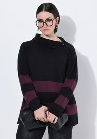 Ung kvinde med kort, mørkt hår iført overdimensionerede firkantede briller, sort og burgunderstribet sweater, holder en sort læder clutch.
