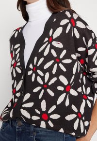 Černý cardigan s velkými bílými sedmikráskami a červenými středy, s designem do V, předními knoflíky a malým logem značky na levé straně.