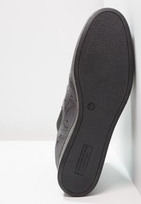 Zapato de ante gris con diseño perforado, suela de goma negra texturizada y marca en relieve en el lateral. Diseño plano y de perfil bajo.