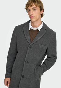 Manteau en mélange de laine gris avec un col cranté, un design à simple boutonnage, deux grands boutons et une poche latérale. Porté sur un pull marron et une chemise blanche.