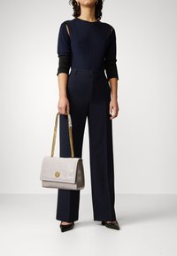 Tuta navy con maniche a taglio, accenti neri e gambe larghe; borsa a mano grigia chiara con motivo in rilievo e tracolla dorata.