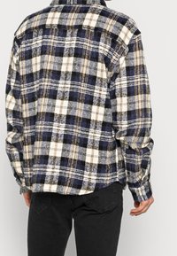 Flanellskjorta med ett mönster av blått, krämfärgat och svart rutmönster, tillverkad av mjukt tyg. Inkluderar långärmar och krage med knäppning.