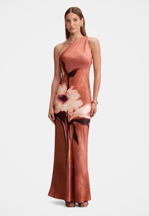 Femme portant une longue robe asymétrique couleur rouille avec un grand imprimé floral blanc et foncé, debout, mains le long du corps, fond neutre.