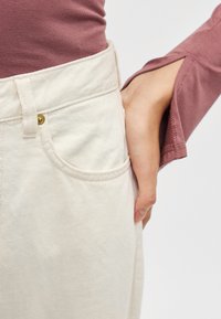 Kreemikas toonis teksapüksid küljetaskuga, millel on kuldse nupuga detail. Seljas burgundia pikkade varrukatega pluus, millel on lõikega aktsendid.