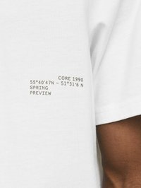Hvid bomulds T-shirt med trykt tekst på ærmet, herunder geografiske koordinater og mærkedetaljer i sort.