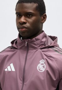 Νέος άνδρας που φοράει μωβ αθλητικό μπουφάν με φερμουάρ και κουκούλα, με τα λογότυπα της Ρεάλ Μαδρίτης και της Adidas στο στήθος.