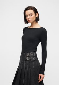 Femme aux cheveux courts et foncés portant un haut noir moulant à manches longues côtelé et une jupe plissée gris foncé à taille haute avec des bretelles à double boucle.