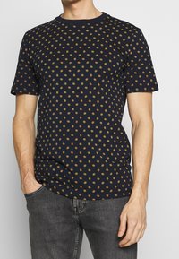 Hombre con camiseta de manga corta azul marino con un pequeño patrón floral amarillo y jeans gris oscuro, con una mano en el bolsillo frente a un fondo neutro.