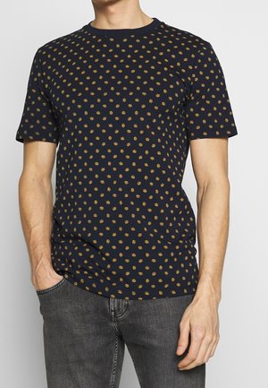Homme portant un t-shirt à manches courtes bleu marine avec un petit motif floral jaune et un jean gris foncé, une main dans la poche, sur un fond neutre.