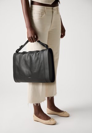 Person in beige cropped pants and cream flats holding a large black leather handbag by its top handle at side.