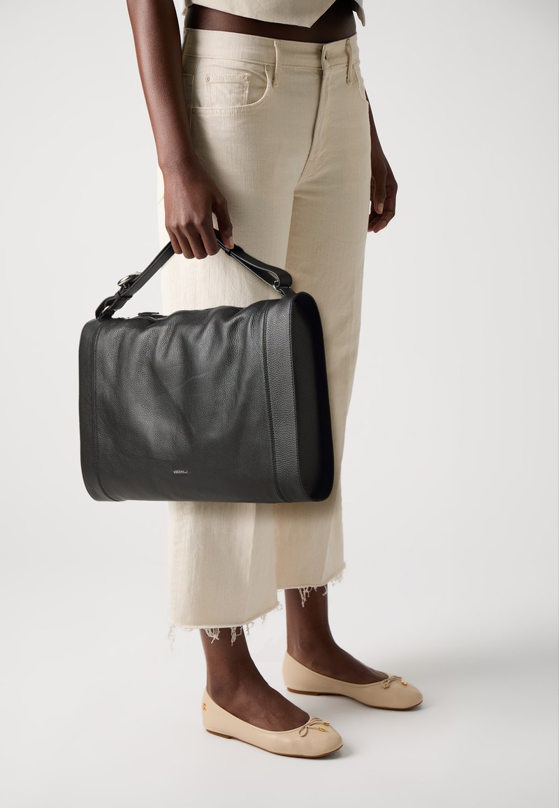 Person in beige cropped pants and cream flats holding a large black leather handbag by its top handle at side.