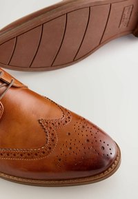 Brązowy skórzany but z perforowanym wzorem wingtips. Posiada okrągły czubek oraz brązową gumową podeszwę z poziomymi rowkami dla lepszej przyczepności.