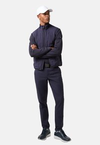 Navy quilted bomber jacket with high collar, paired with slim-fit navy pants and navy sneakers, complemented by a white cap.
