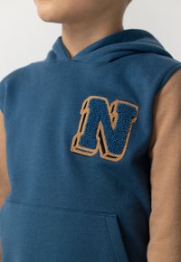 Bambino che indossa un hoodie senza maniche blu con un grande patch testurizzato a forma di "N" sul petto e maniche lunghe marroni sotto.