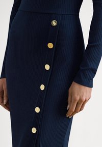 Marineblå ribbestrikket kjole med lange ermer og høyt splitt på låret, med gullfargede dekorative knapper langs siden som detalj.