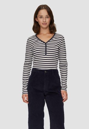 Chemise rayée à manches longues en noir et blanc, avec un col en V et une patte de boutonnage. Associée à un pantalon en velours côtelé bleu marine foncé.