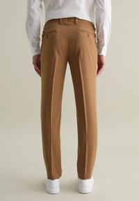 Pantalones marrones a medida con una textura suave, que presentan dos bolsillos traseros con botón y se llevan con zapatillas blancas.