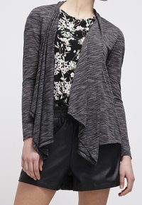 Cardigan grigio leggero con apertura frontale e design drappeggiato indossato sopra una maglia nera a fantasia floreale, abbinato a pantaloni corti neri.